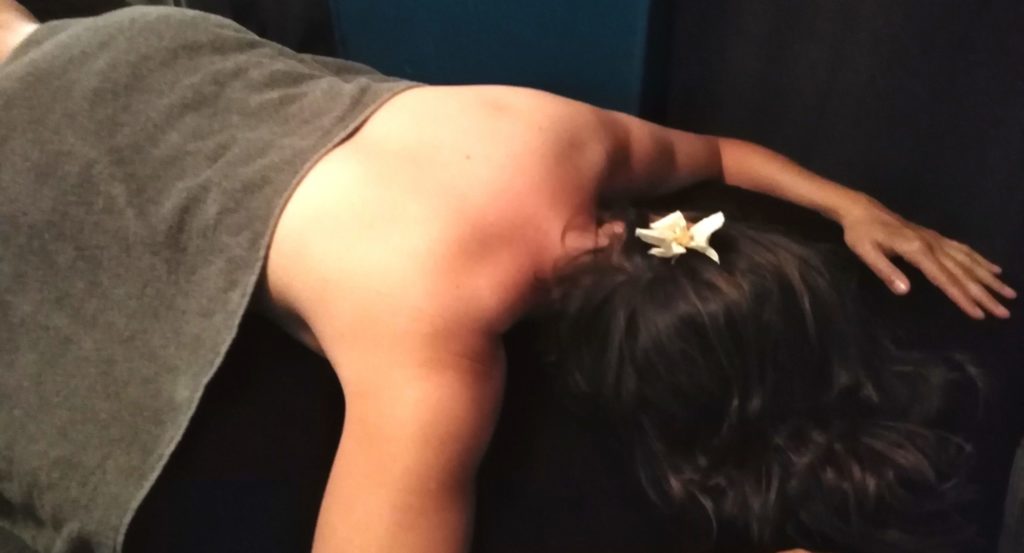 Jeune femme allongée sur le ventre, une fleur dans les cheveux. Elle est dans la salle de soins, allongée sur la table de massage drapée de noir, une serviette grise posée sur le bas du dos.