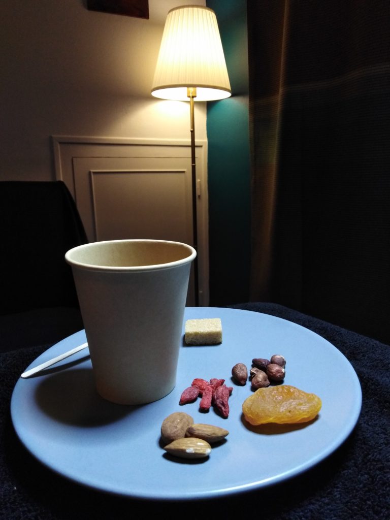 Intérieur cosy de la salle de soins.Collation sur une assiette à dessert bleue. Tisane dans un gobelet en carton beige,fruits secs (abricot, amandes, noisettes, baies de gogi). Une lampe sur pied allumée dans le coin de la pièce met en valeur les boiseries peintes.