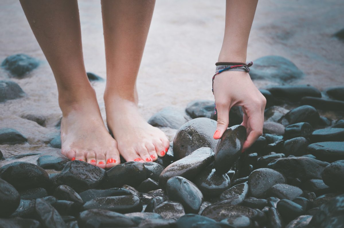 Pieds et mains d'une jeune femme qui se penche pour ramasser un galet sur une plage.