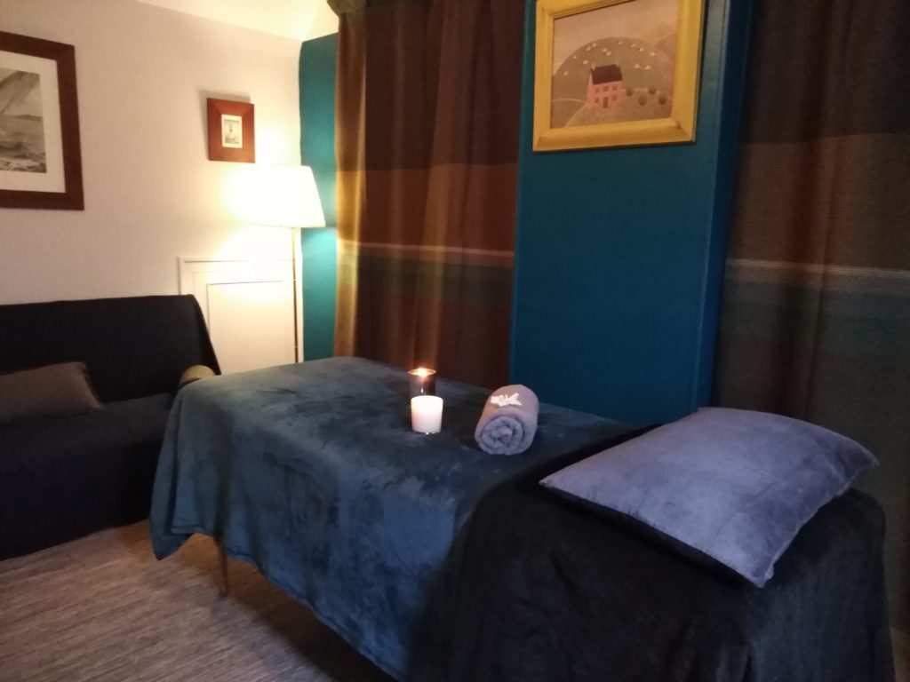 Institut de beauté bio. Photo de la salle de soins présentant une table de massage sur laquelle sont posées des bougies allumées. Des cadres sont accrochés aux mur. La salle dispose d'un canapé et de deux fenêtres occultées par d'épais rideaux. C'est un intérieur cosy!
